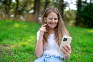 A woman smiles while using her smartphone outdoors in a lush green park on a sunny day, enjoying her time in nature