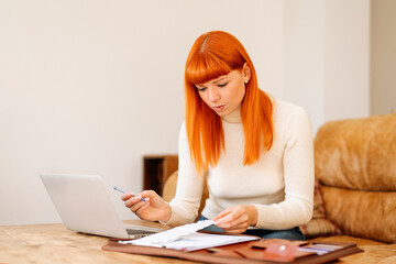 Young woman with orange hair working at home, organizing documents on a wooden desk during daytime...
