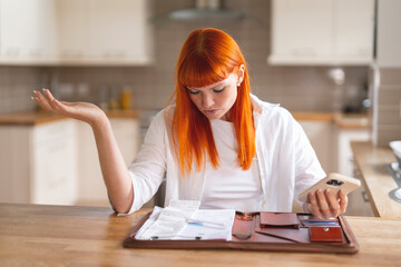 Young woman reviews financial documents and phone messages in modern kitchen, feeling stressed and...