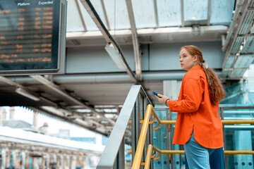 A woman in an orange shirt checks her phone on a train platform during a busy afternoon in a city
