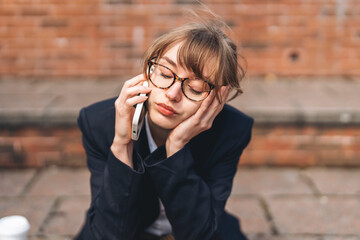 Young woman in business attire sitting outside, thoughtfully talking on the phone while resting her head on her hand