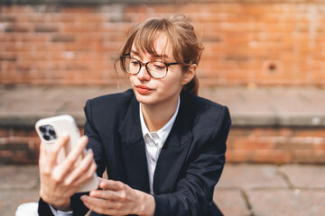 Young woman in formal attire checks her smartphone while sitting on steps in a city, enjoying a sunny day outdoors