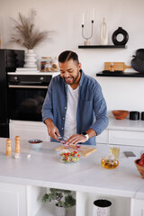 Young smiling african american man cutting onion for fresh salad near smartphone and tea at home
