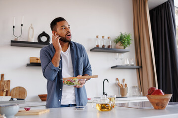 African american man using wireless earphones and holding fresh salad in kitchen at home