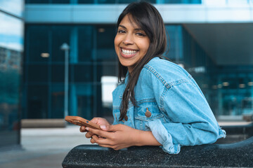 Young woman happily using her smartphone while sitting outside a modern building on a sunny day
