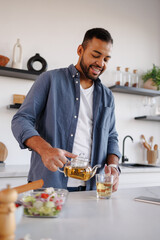 Smiling black man pouring herbal tea near smartphone and vegetable salad in kitchen