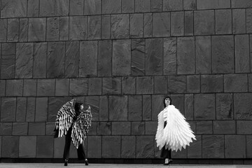 Two models showcase striking feather winged outfits against a textured dark wall in a fashion...