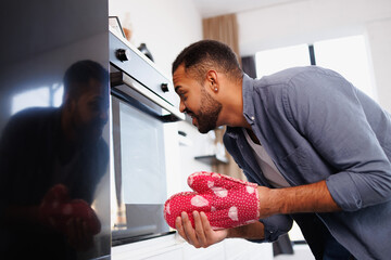 Bearded african american man taking off glove and looking at oven in kitchen at home