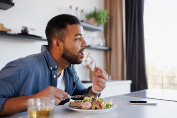 Bearded african american man eating fresh salad and chicken legs near smartphone and blurred tea in...