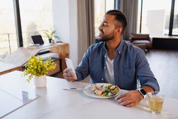 Smiling african american man holding smartphone near fresh salad, chicken legs and tea at home