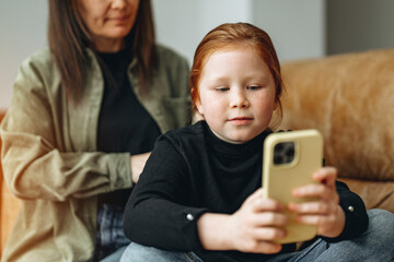 A young child focused on a smartphone while sitting on a couch with a family member in a cozy living room setting