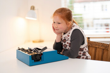 A young girl admires a vintage typewriter while sitting at a bright table by the window in her home...