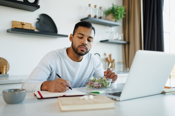 Fototapeta premium African american freelancer writing on notebook near fresh salad and laptop at home