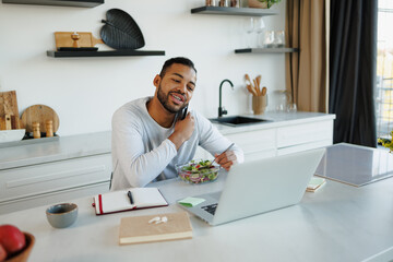 Smiling african american freelancer talking on smartphone near laptop and salad in kitchen