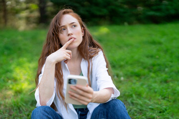 Young woman sits on grass in a park and thoughtfully uses her smartphone while deep in...