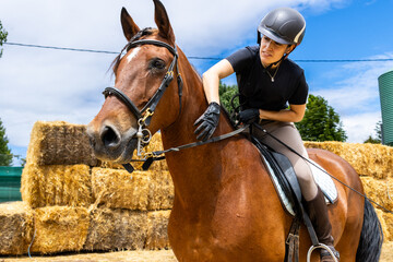 Female rider adjusting reins on a brown horse near hay bales - Powered by Adobe