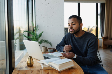 African american freelancer holding smartphone near laptop and book at home