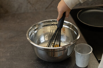 A small hand holds a black whisk and makes pancake batter in a silver bowl