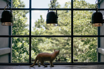 A view of a large square glass window decorated with a lamp. A cat doll stands looking out the window with green leaves in the background