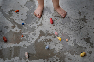 Multicolored chalks scattered on concrete floor with puddles, little child's dirty bare feet