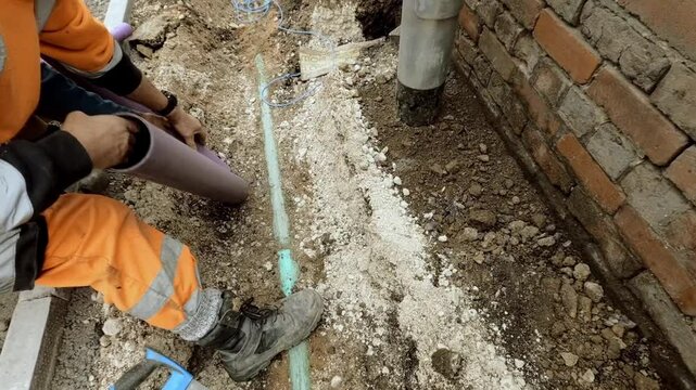 Worker installing a telecom duct fro internet broadband cable  alongside a brick wall in a construction site during the day