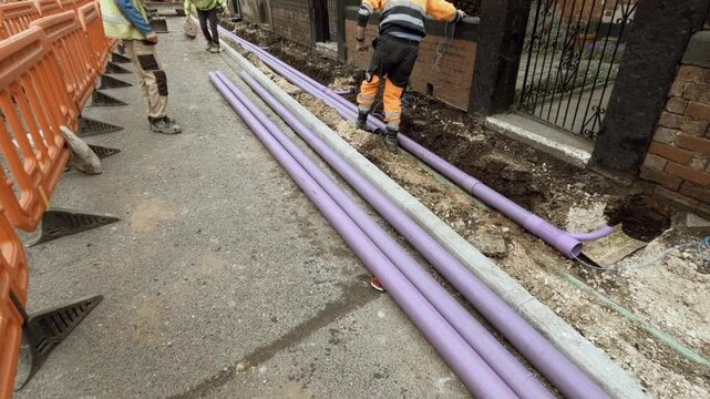 Worker installing a telecom duct fro internet broadband cable  alongside a brick wall in a construction site during the day