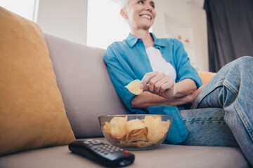 Cropped portrait of nice aged woman hold eat chips watch tv wear blue shirt bright interior flat...
