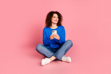 Young woman casual outfit sitting with a smartphone against a pink background, enjoying her mobile experience.