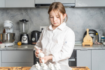 A cute little girl pastry chef in a chef's robe holds a pastry bag in her hands and squeezes meringue onto baking paper.