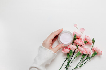 Under eye cream bottle in woman hand with pink flowers on wooden white background. Delicate cream for skincare