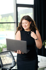 A talented young woman with long curly hair engages with her laptop, embodying confidence in a vibrant corporate environment.