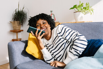 Happy young Latin American woman relaxing on sofa sending a voice note audio message on mobile phone while enjoying cozy winter day at home
