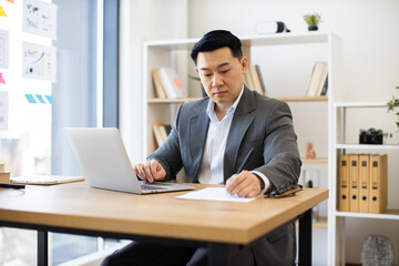 Obraz premium Asian businessman in modern office focused on laptop while writing report. Wearing formal attire, mid-aged male demonstrates concentration in professional environment.