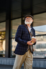 A fashionable senior man strolls confidently near a contemporary building, savoring a warm beverage in autumn.