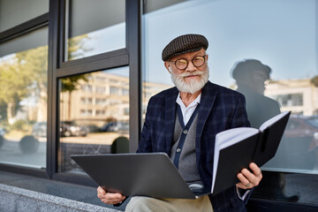 A dapper senior man with a laptop sitting near a contemporary building amid autumn colors.