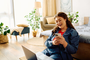 A young plus-size woman with curly hair smiles brightly as she enjoys time at home, engaged with her smartphone and laptop.