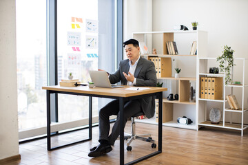 Asian male businessman in his 30s engaged in online video call while working on laptop in modern office. Professional environment with charts on wall and organized workspace.