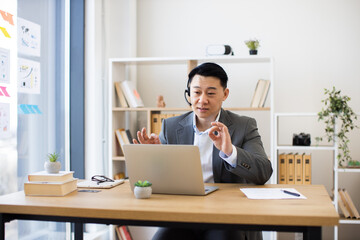 Asian businessman in his 30s wearing headset during online video call in modern office. He sits at desk with laptop, gesturing while speaking.