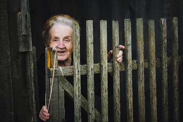 A portrait of an elderly woman peeking out from behind a wooden village fence, her eyes filled with curiosity and warmth.