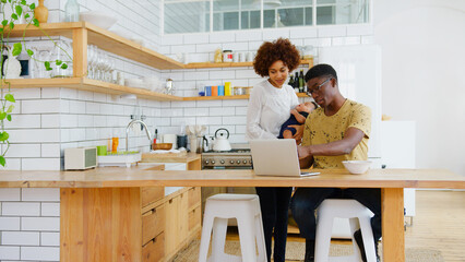 Fototapeta premium Busy Family In Kitchen At Breakfast With Father Working On Laptop And Mother Caring For Baby Son