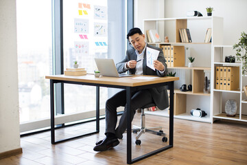 Asian businessman wearing headset in office showing graphs during online video call. Professional setting with laptop, modern interior, and large window. Business attire suggests formal meeting.