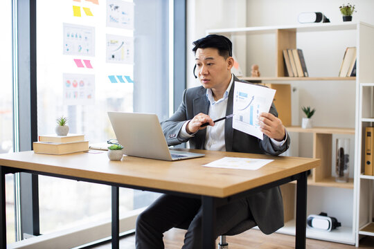 Portrait of Asian male professional in mid-30s wearing headset during online call. He presents detailed report with graphs and charts while sitting at modern office desk.