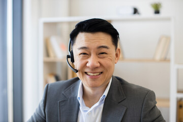 Asian male businessman in office wearing headset and smiling during online video call. Professional atmosphere with bookshelves in background.