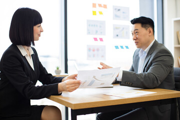 Caucasian businesswoman shows report to Asian businessman in office setting. Professionals engage in discussion over printed documents, emphasizing teamwork and collaboration.