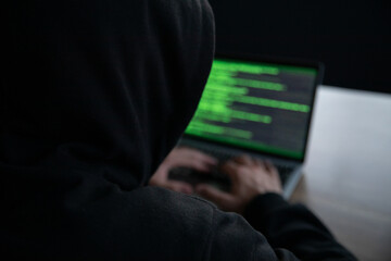 A person in a dark hoodie types on a laptop displaying green code. The image represents themes of hacking, cybersecurity, digital security, and online anonymity in a low-light environment.