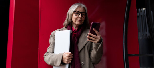 A senior businesswoman standing outside, checking her smartphone while holding a portfolio on a vibrant red background
