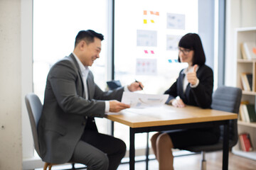 Blurred background. Asian businessman presents report to Caucasian businesswoman in office setting. Both individuals display positive and professional demeanor.