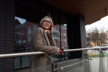 Middle-aged businesswoman stands confidently on balcony in urban setting during late afternoon, reflecting on career achievements