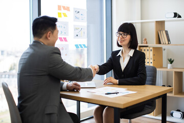 Caucasian businesswoman and Asian businessman shaking hands in modern office setting. Business professionals reaching agreement, sealing contract, partnership success symbolized.