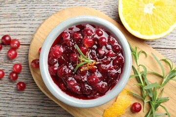 Tasty cranberry sauce in bowl, berries, orange and rosemary on wooden table, flat lay